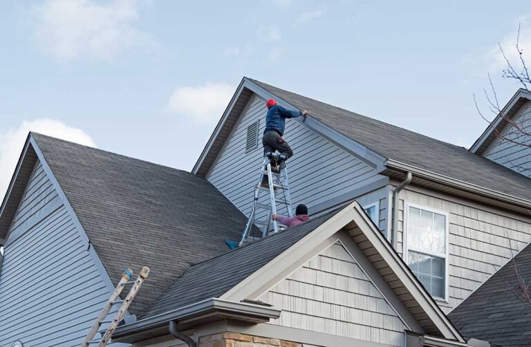Person clearing leaves from a residential gutter beside a shingled roof with a downspout below.