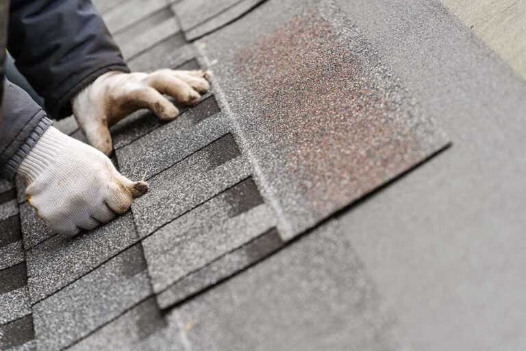 Homeowner examining curled shingles, rusted flashing, and roof granules beside a ladder after winter weather.